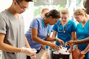 Multi-ethnic group of College students in nursing class. Woman professor is explaining how to do blood tests. Everybody is wearing a uniform. They are in a mock hospital room with mannequin arm on table. Horizontal indoors waist up shot.